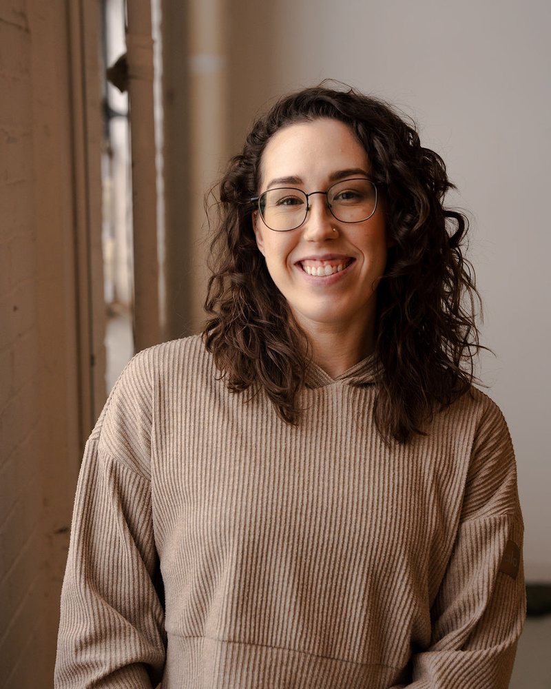 Kristina smiling at the camera in a soft beige sweater, standing in natural window light.